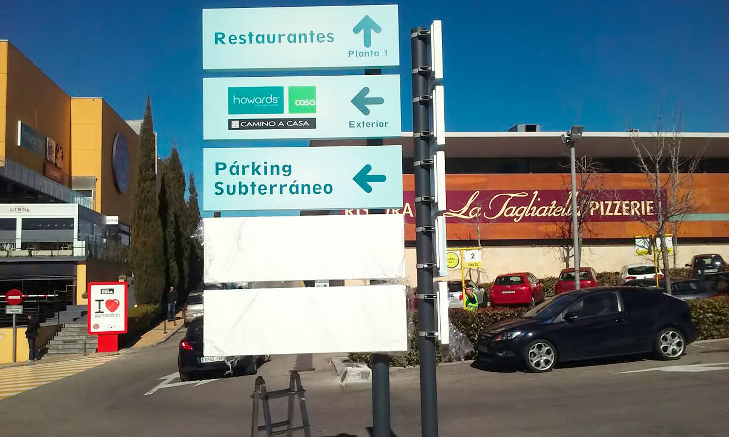 Poste de señalización con carteles direccionales en un área comercial, indicando la ubicación de restaurantes, tiendas y el aparcamiento subterráneo.